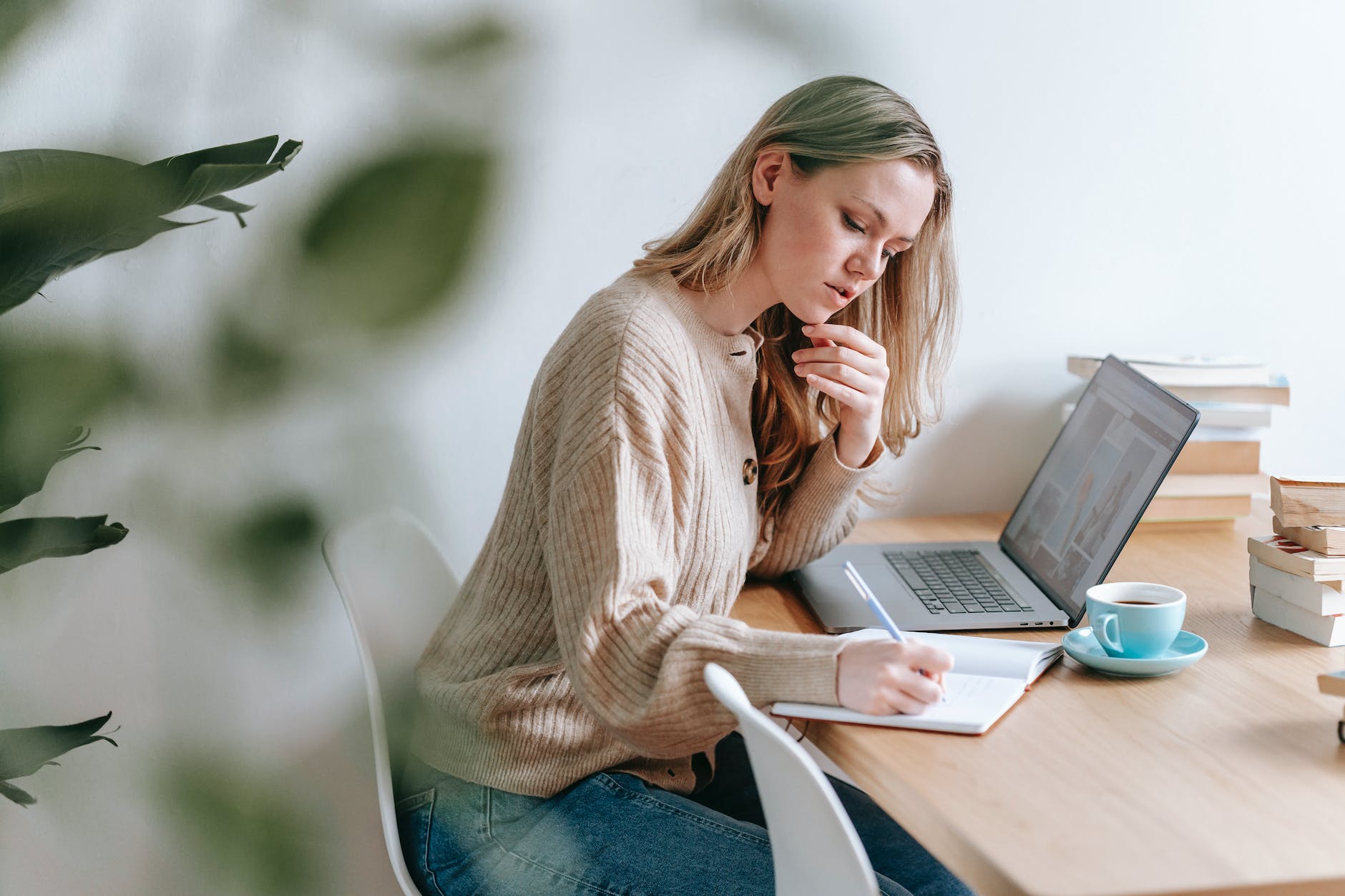 pensive woman taking notes at table with laptop and cup of coffee